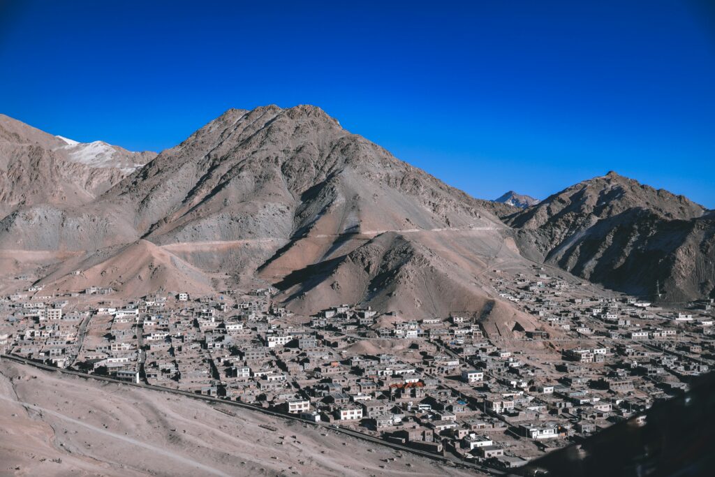 A stunning aerial view of Leh with its rugged mountain backdrop and scattered village buildings under a clear blue sky.