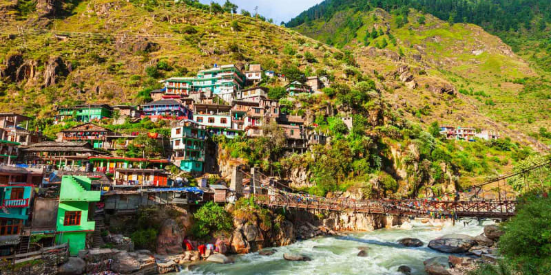small Himalayan village beside a clear mountain river, wooden guesthouses and cafes along the bank, Kasol village beside the Parvati River on a day trip from Manali