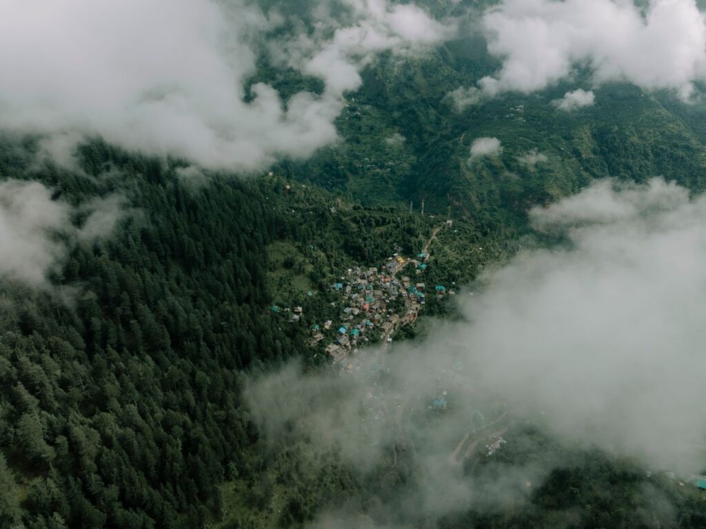 Aerial photograph showcasing a small village nestled among lush green mountains and clouds in Himachal Pradesh, India.