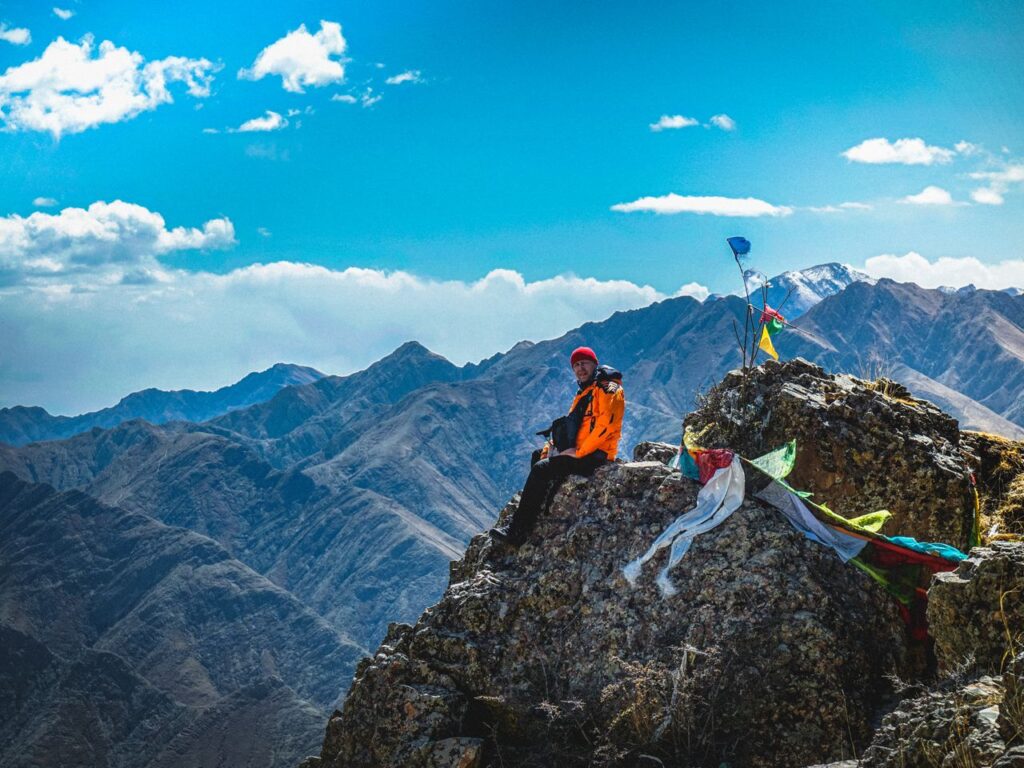 Full body of hiker in warm clothing and hat looking at camera while resting on cliff in ravine under light blue sky in sunlight Traveller acclimatising during a Langza village Spiti high-altitude day trip