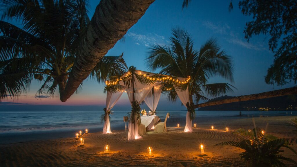 Late-afternoon scene at a Port Blair beach with a small beachside restaurant under palm trees, simple tables, soft golden light, calm sea behind. A few diners seen only from a distance. No readable signage, Beachside refreshments near Corbyn's Cove Beach at golden hour