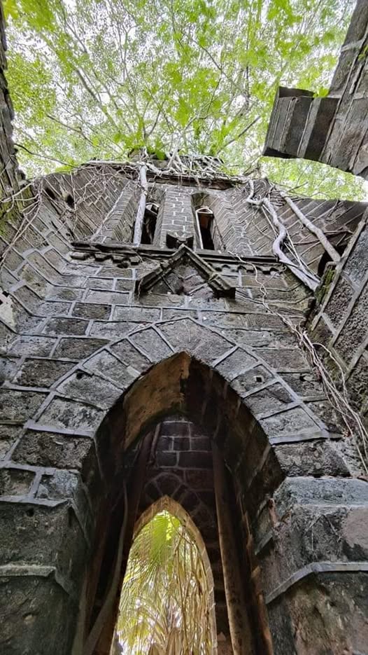British-era church on Ross Island, Andaman. Weathered stone arches and walls with thick banyan roots growing over them. Soft green light filtering through tall trees, a narrow shaded path in the foreground. Calm, quiet mood Banyan roots over old church ruins on Ross Island Andaman