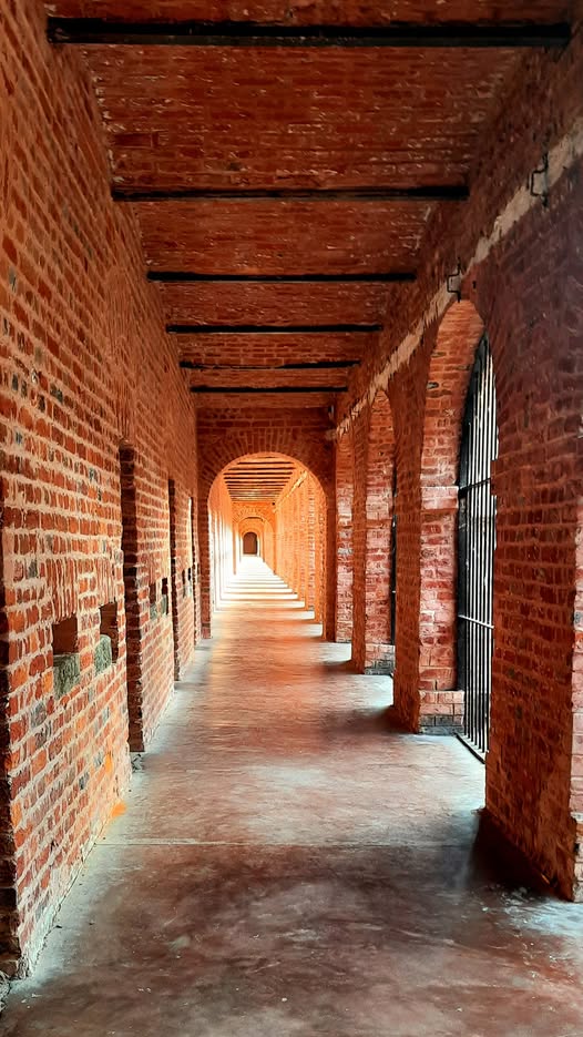 Cellular Jail wing in Port Blair. Rows of old iron-grilled cell doors on one side, soft daylight from the open side, long perspective, quiet and solemn mood Empty corridor with iron-grilled cell doors inside the Cellular Jail in Port Blair