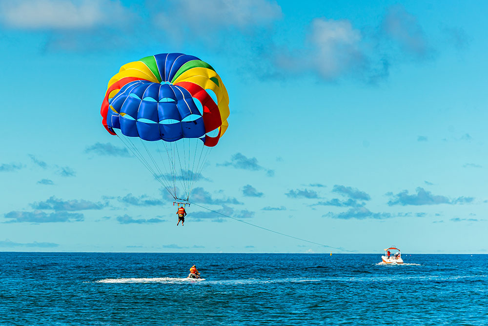 parasail high above a calm bay near a palm-fringed beach in Port Blair, Andaman. A speedboat is visible below, small in the frame. Bright daylight, blue sea, tropical atmosphere. Parasailing above the water at Corbyn's Cove Beach near Port Blair